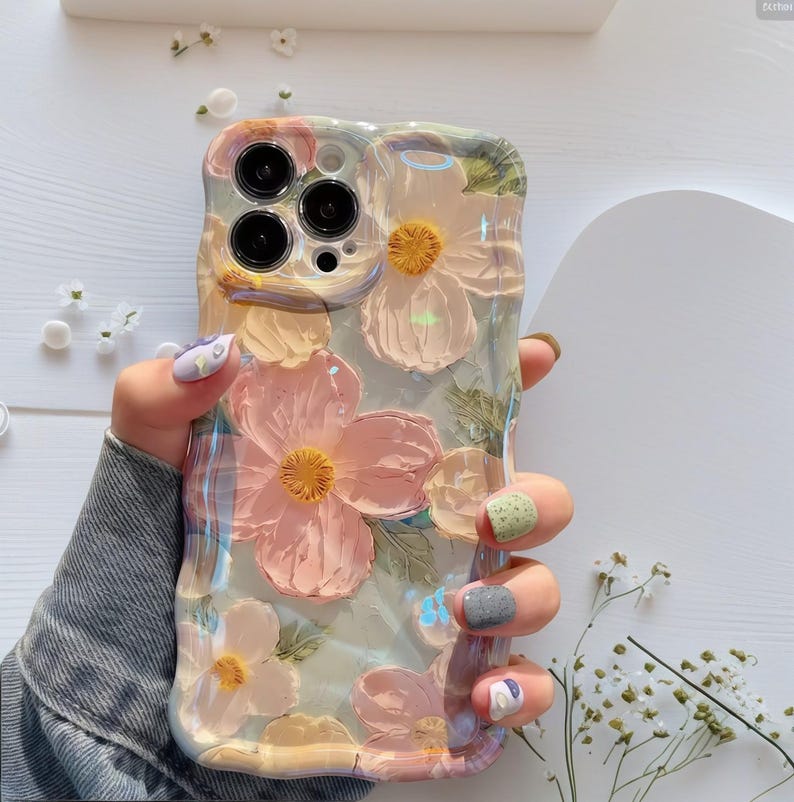 Pode incluir: Capa de telefone transparente com um design floral. A capa apresenta flores rosa e amarelas texturizadas em relevo com folhas verdes. A capa tem uma borda ondulada e &eacute; segurada na m&atilde;o.