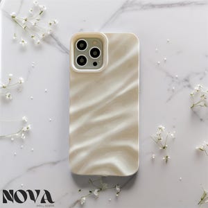 Peut inclure: Coque de téléphone blanc nacré avec un motif ondulé, avec une découpe pour l'appareil photo. L'étui est posé sur un fond en marbre avec de délicates fleurs blanches. La marque "NOVA SHELL DESIGN" est visible en bas.