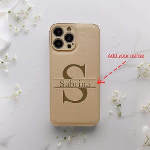 Peut inclure: Coque de téléphone beige avec une grande lettre "S" et le nom "Sabrina" gravés. L'étui a une surface texturée et une découpe pour l'appareil photo. Le texte "Add your name" est écrit en rouge.