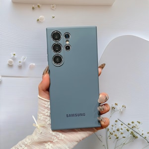 Op de afbeelding: Een leisteenblauwe Samsung-telefoon met meerdere cameralenzen wordt in een hand gehouden. De achterkant van de telefoon heeft het Samsung-logo. De hand draagt een kanten handschoen. Kleine witte bloemen en parels zijn rond de telefoon verspreid.