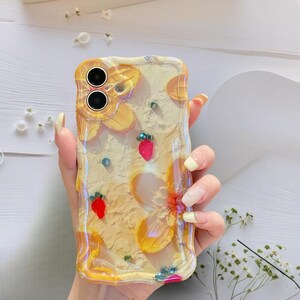 May include: Clear phone case with a yellow floral design and embedded red strawberries. The case has a textured surface and a wavy edge. Small blue and green embellishments are scattered throughout the design.