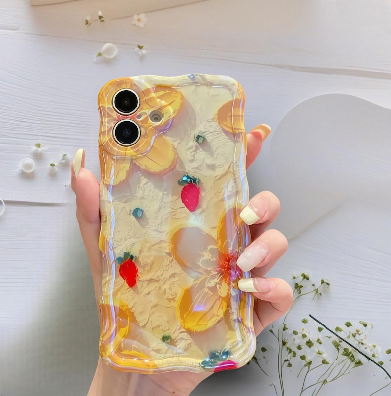 May include: Clear phone case with a yellow floral design and embedded red strawberries. The case has a textured surface and a wavy edge. Small blue and green embellishments are scattered throughout the design.