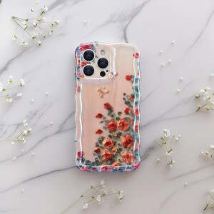 Pode incluir: Uma capa de telefone transparente com um design floral. A capa apresenta um padr&atilde;o de rosas vermelhas e laranja com folhas verdes. As bordas t&ecirc;m uma borda ondulada e iridescente. As lentes da c&acirc;mera do telefone s&atilde;o vis&iacute;veis.