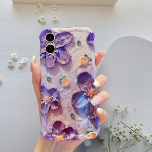 May include: A clear phone case with a textured, off-white background and purple floral designs. The case features raised, three-dimensional flowers and small orange fruit accents. The case is held in a hand with yellow nail polish.