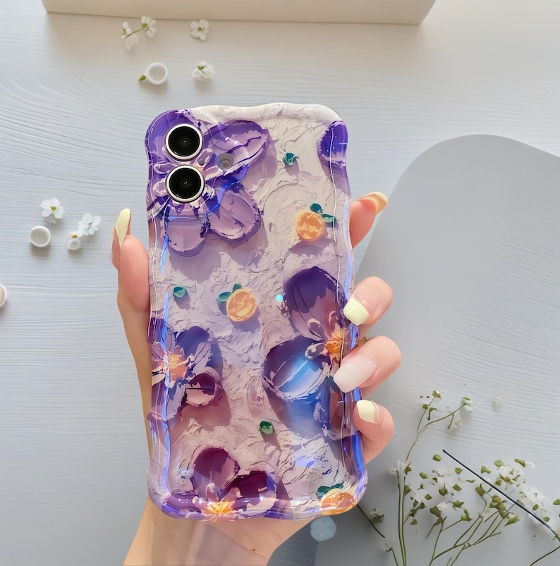 May include: A clear phone case with a textured, off-white background and purple floral designs. The case features raised, three-dimensional flowers and small orange fruit accents. The case is held in a hand with yellow nail polish.