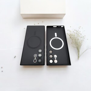 Może przedstawiać: Dwa czarne etui na telefon o minimalistycznym designie. Jedno etui jest otwarte, odsłaniając biały okrągły kontur, a drugie zamknięte. Oba etui mają okrągłe wycięcia na aparat i duży okrągły obszar ładowania magnetycznego.
