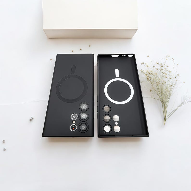 Może przedstawiać: Dwa czarne etui na telefon o minimalistycznym designie. Jedno etui jest otwarte, odsłaniając biały okrągły kontur, a drugie zamknięte. Oba etui mają okrągłe wycięcia na aparat i duży okrągły obszar ładowania magnetycznego.