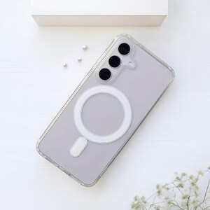 Peut inclure: Coque de téléphone transparente avec un anneau MagSafe blanc et un petit rectangle blanc en bas. La coque a un dos gris et un bord transparent. Le téléphone a trois objectifs de caméra noirs. La coque est sur une surface blanche.