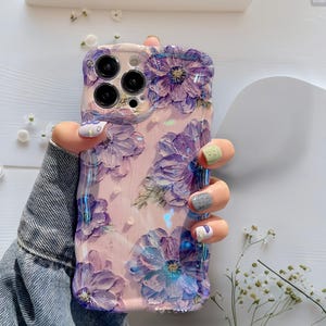 Pode incluir: Uma capa de telefone com um design floral. A capa &eacute; de cor rosa claro e apresenta flores roxas em relevo com detalhes azuis. A capa tem um acabamento brilhante. A capa do telefone &eacute; segurada por uma pessoa que usa uma jaqueta jeans.