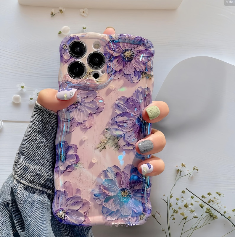 Pode incluir: Uma capa de telefone com um design floral. A capa &eacute; de cor rosa claro e apresenta flores roxas em relevo com detalhes azuis. A capa tem um acabamento brilhante. A capa do telefone &eacute; segurada por uma pessoa que usa uma jaqueta jeans.