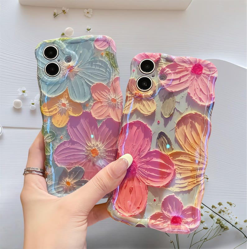 May include: Two phone cases with a floral design. The cases feature raised, textured flowers in shades of pink, purple, orange, and blue. The cases have a glossy finish and are held up by a person's hand.