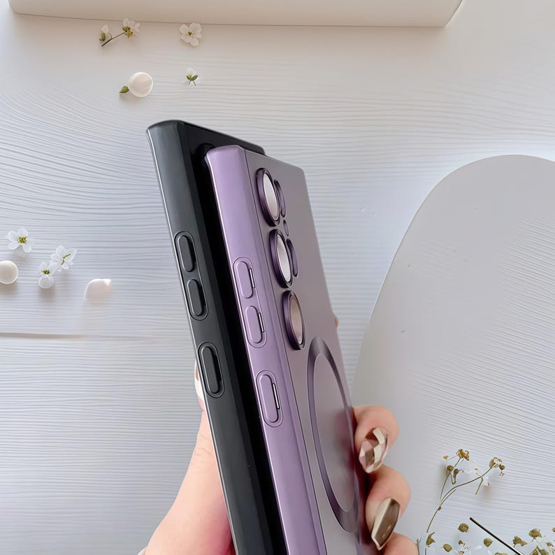 Może przedstawiać: Dwa etui na telefon, jedno czarne i jedno lawendowe, są ułożone jedno na drugim. Lawendowe etui ma okrągły element i wycięcia na aparat. Czarne etui jest na dole, a oba mają wycięcia na przyciski. Tło jest jasne i neutralne.