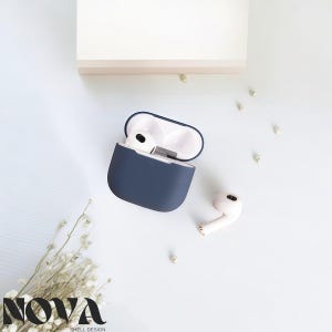 May include: A navy blue protective case for wireless earbuds, open to reveal the white earbuds inside. One earbud rests outside the case. The case has a white interior and a dark blue exterior. The word "NOVA" is printed in black at the bottom of the image.