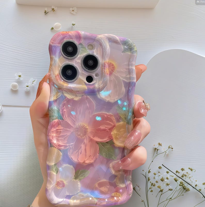 Pode incluir: Uma capa de telefone com um design floral e acabamento perolado. A capa apresenta flores tridimensionais em relevo em tons de rosa, p&ecirc;ssego e branco sobre um fundo lavanda. A capa tem um brilho brilhante e iridescente e um recorte para a c&acirc;mera.