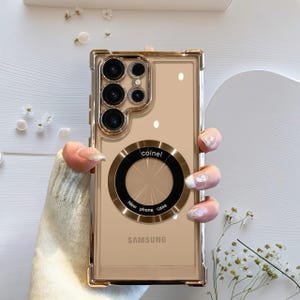 Può includere: Una custodia trasparente per telefono con una cornice color oro e un supporto ad anello circolare. La custodia ha un retro color oro con il logo Samsung e la scritta "New phone case" attorno al supporto ad anello. La custodia è tenuta da una persona.