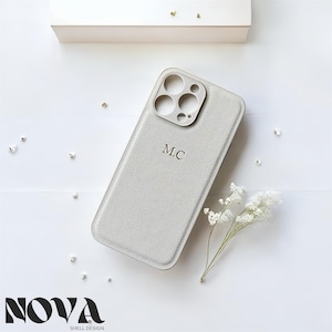Peut inclure: Un étui de téléphone gris clair avec les initiales "M.C." est présenté. L'étui a une surface texturée et une découpe pour l'appareil photo. Le logo "NOVA SHELL DESIGN" est en bas. De petites fleurs blanches et des perles décorent l'image.
