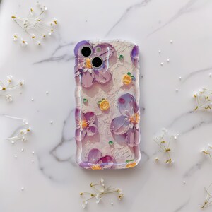May include: Clear phone case with a floral design. The case features purple flowers and small yellow accents on a textured background. The case is designed to fit a smartphone.