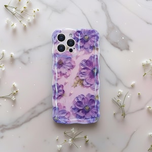 Pode incluir: Uma capa de telefone transparente com uma borda ondulada, apresentando um design floral em aquarela de flores roxas em um fundo rosa claro. A capa tem um acabamento brilhante e &eacute; decorada com pequenas flores brancas.