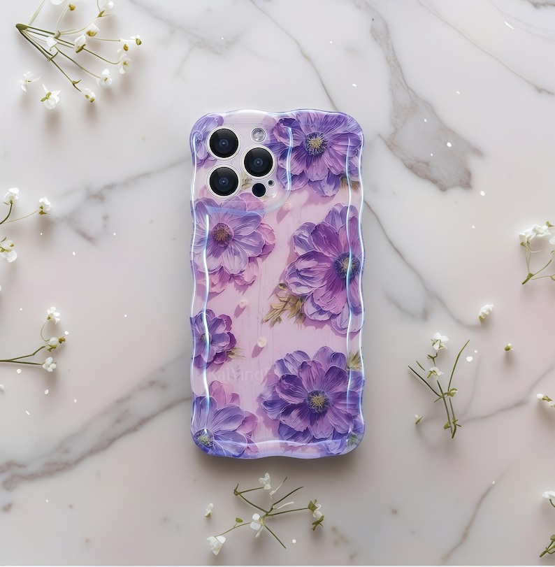 Pode incluir: Uma capa de telefone transparente com uma borda ondulada, apresentando um design floral em aquarela de flores roxas em um fundo rosa claro. A capa tem um acabamento brilhante e &eacute; decorada com pequenas flores brancas.