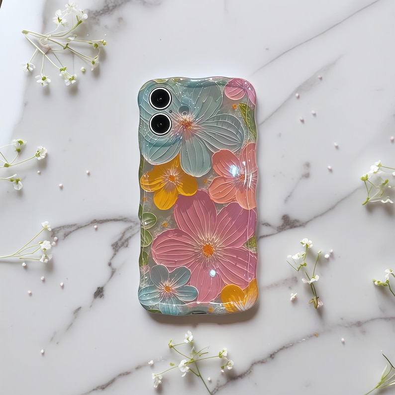 May include: A clear phone case with a floral design. The case features embossed flowers in shades of pink, yellow, and blue. The case has a textured, wavy edge. The phone case is on a white marble surface.
