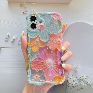 May include: A clear phone case with a floral design. The case features embossed flowers in shades of pink, orange, yellow, and blue. The case has a wavy edge and is held in a hand.
