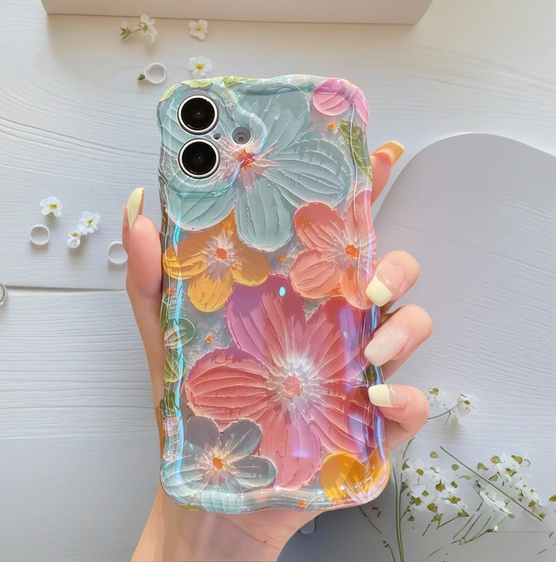 May include: A clear phone case with a floral design. The case features embossed flowers in shades of pink, orange, yellow, and blue. The case has a wavy edge and is held in a hand.