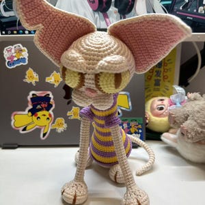 May include: A handmade crochet creature with large, pink-edged ears and yellow-green eyes. The creature has a beige head and limbs, with a purple and yellow striped shirt. The tail is curled, and the feet are detailed.
