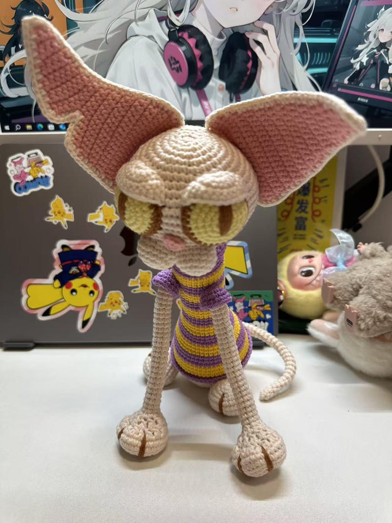 May include: A handmade crochet creature with large, pink-edged ears and yellow-green eyes. The creature has a beige head and limbs, with a purple and yellow striped shirt. The tail is curled, and the feet are detailed.