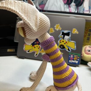 May include: Crocheted animal figurine in cream, yellow, and purple. The handmade toy features a long neck with yellow and purple stripes, long legs, and a large head with floppy ears. The toy is sitting on a white surface.