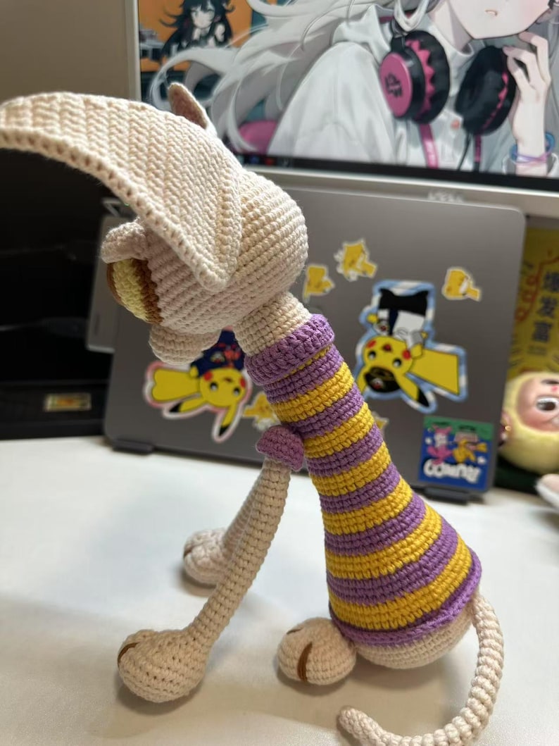 May include: Crocheted animal figurine in cream, yellow, and purple. The handmade toy features a long neck with yellow and purple stripes, long legs, and a large head with floppy ears. The toy is sitting on a white surface.