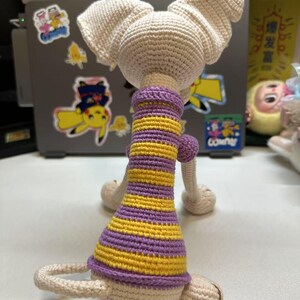 May include: A handmade crochet doll in a seated position, viewed from the back. The doll has large, pointed ears and is wearing a purple and yellow striped dress. The doll is made of cream-colored yarn.