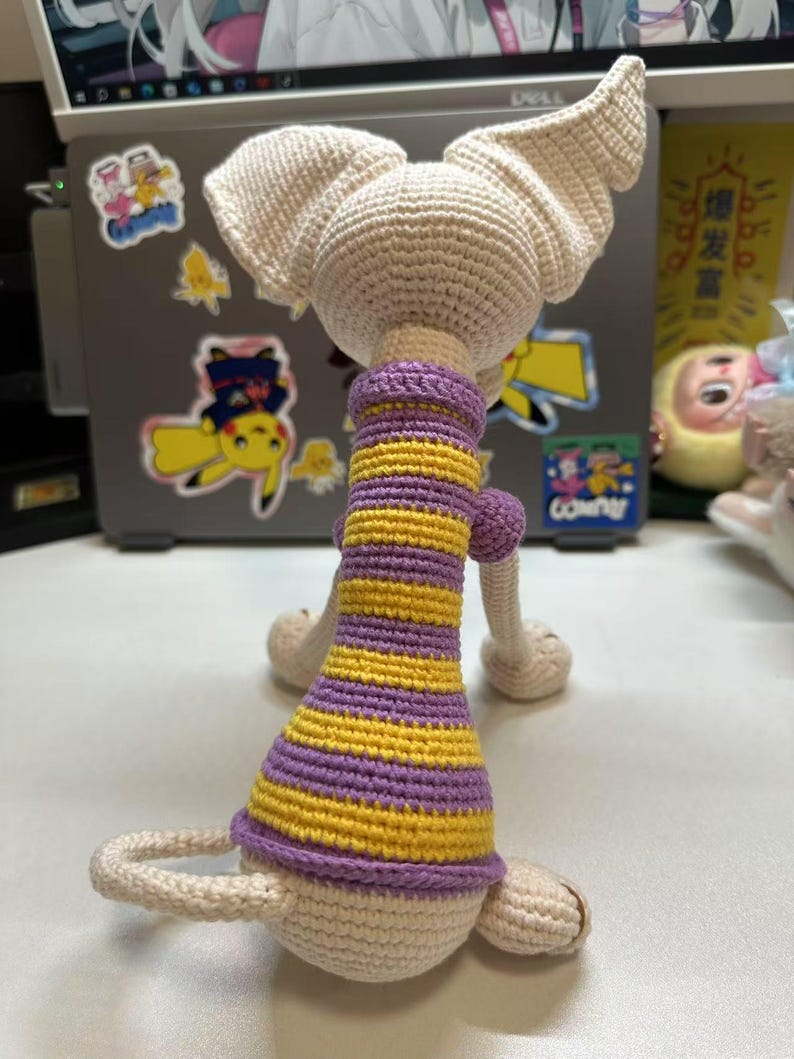 May include: A handmade crochet doll in a seated position, viewed from the back. The doll has large, pointed ears and is wearing a purple and yellow striped dress. The doll is made of cream-colored yarn.