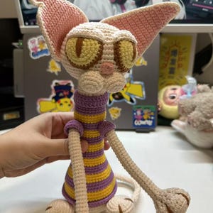 May include: A handmade crochet cat doll with large ears and expressive yellow eyes. The cat wears a purple and yellow striped shirt and has long, slender limbs. The cat is made of beige, pink, and brown yarn.