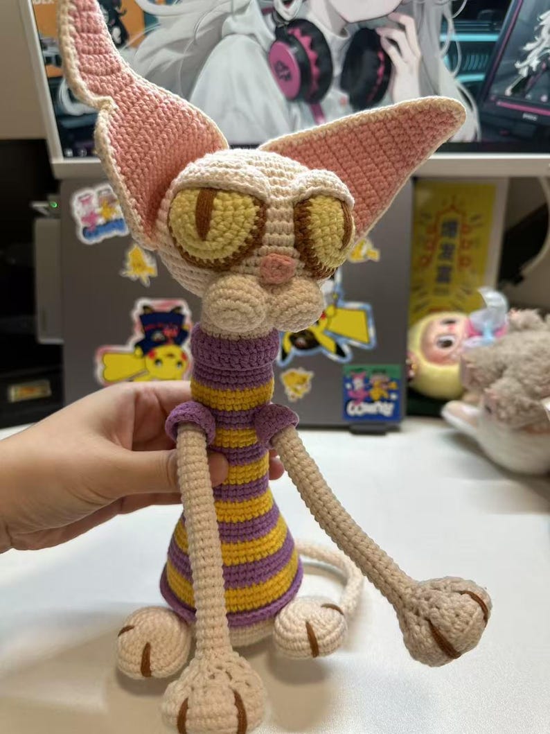 May include: A handmade crochet cat doll with large ears and expressive yellow eyes. The cat wears a purple and yellow striped shirt and has long, slender limbs. The cat is made of beige, pink, and brown yarn.