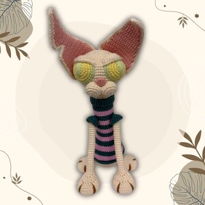 May include: A handmade crochet cat doll with large, expressive yellow and green eyes, pink ears, and a striped neck. The cat has long legs and feet with brown details. The doll is made with cream, pink, green, and purple yarn.