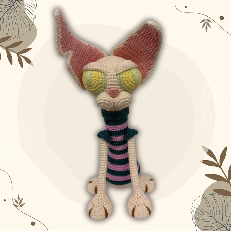 May include: A handmade crochet cat doll with large, expressive yellow and green eyes, pink ears, and a striped neck. The cat has long legs and feet with brown details. The doll is made with cream, pink, green, and purple yarn.