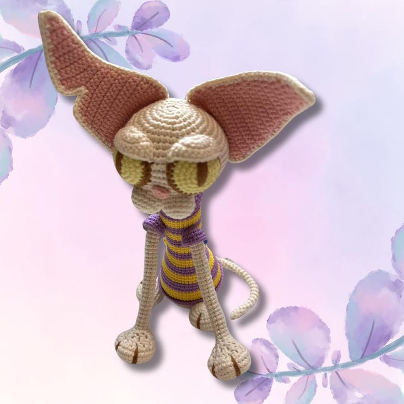 May include: A handmade crochet cat doll with large ears and yellow eyes. The cat wears a purple and yellow striped shirt. The doll is beige, with pink accents on the ears and nose. The cat is sitting down.