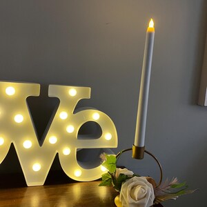 Puede incluir: Una composición decorativa con una vela LED blanca en un soporte dorado, con una rosa blanca y vegetación. La palabra "love" está iluminada con luces.