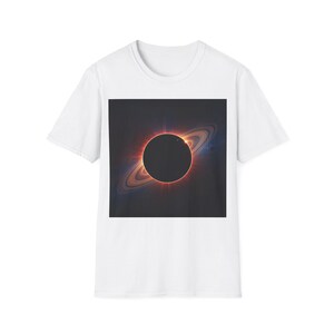 Könnte beinhalten: Ein weißes T-Shirt mit kurzen Ärmeln und einem mittigen, quadratischen Aufdruck einer totalen Sonnenfinsternis. Das Motiv zeigt eine dunkle kreisförmige Silhouette, umgeben von einer leuchtend orangefarbenen Korona und Planetenringen.
