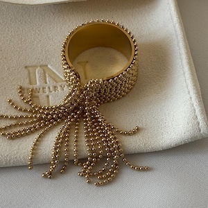 Può includere: Anello dorato con texture a perline e design a nappe. L'anello è su una superficie bianca e la nappa è composta da piccole perline pendenti. L'anello sembra essere un accessorio di moda.
