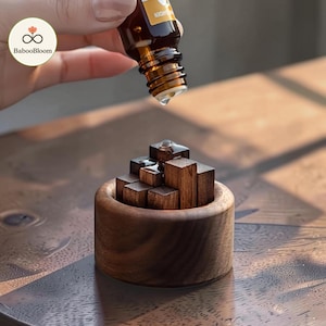 May include: A small, round, dark brown wooden bowl filled with several vertical wooden blocks of varying heights. A clear liquid drop falls from an amber glass dropper bottle into the center of the wooden blocks, which are arranged in a cluster.