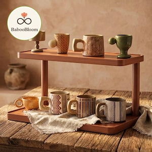 May include: A wooden display shelf holds a collection of ceramic mugs and a small decorative cup. The mugs feature various textures and designs, including geometric patterns and earthy tones. The shelf is on a rustic wooden table.