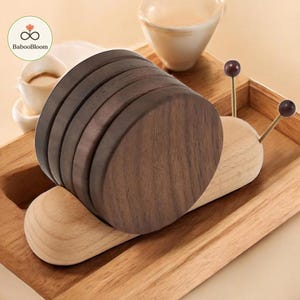 May include: A wooden snail-shaped holder containing five circular dark brown coasters. The snail body is light-colored wood with two thin metal antennae topped with dark brown spheres. The coasters are stacked vertically to form the snail shell.