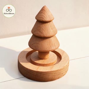 Puede incluir: Una figura de árbol de madera de color marrón claro con una parte superior cónica de tres niveles montada sobre un tronco cilíndrico. El árbol se asienta sobre una base circular de madera con un centro rebajado, mostrando veta natural.