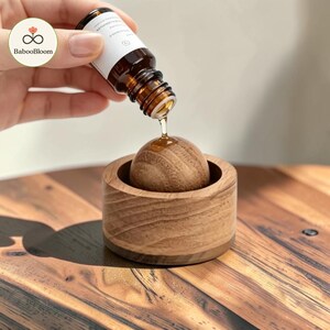May include: A small amber glass dropper bottle with a white label is dispensing a clear liquid onto a smooth wooden sphere that is nested inside a matching circular wooden base, showcasing the natural grain and texture of the light brown wood.