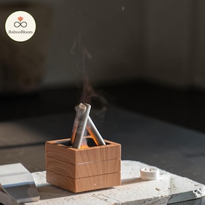 Puede incluir: Un difusor cuadrado de grano de madera claro con una abertura central y dos componentes inclinados en forma de palo. Un cálido resplandor naranja emana del centro, acompañado por una fina columna de vapor ascendente. El logo BabooBloom es visible.