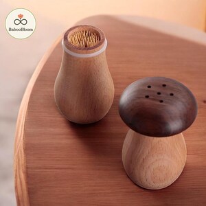 May include: Wooden mushroom-shaped toothpick holder and salt shaker set. The toothpick holder is light brown with a white rim and filled with toothpicks. The salt shaker has a dark brown top with six holes.