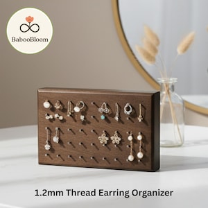 Może przedstawiać: Ciemnobrązowy drewniany organizer na kolczyki z wieloma małymi kołkami do zawieszania kolczyków. Organizer jest prostokątny i zawiera różne kolczyki. Tekst "1.2mm Thread Earring Organizer" znajduje się na dole.