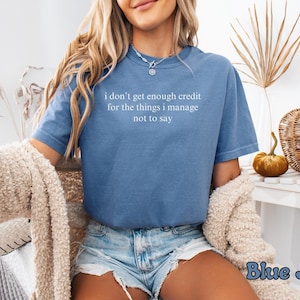 K&ouml;nnte beinhalten: Blaues Jeans-T-Shirt mit dem wei&szlig;en Text "i don't get enough credit for the things i manage not to say". Das Shirt wird mit Jeansshorts kombiniert. Ein beiger Pullover ist &uuml;ber den Schultern drapiert.
