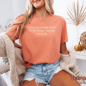 K&ouml;nnte beinhalten: Terracottafarbenes T-Shirt mit dem wei&szlig;en Text "I don't get enough credit for the things I manage not to say". Das Shirt wird mit Jeansshorts und einer cremefarbenen Strickjacke kombiniert. Dekorative K&uuml;rbisse und ein Weidenkorb sind im Hintergrund.
